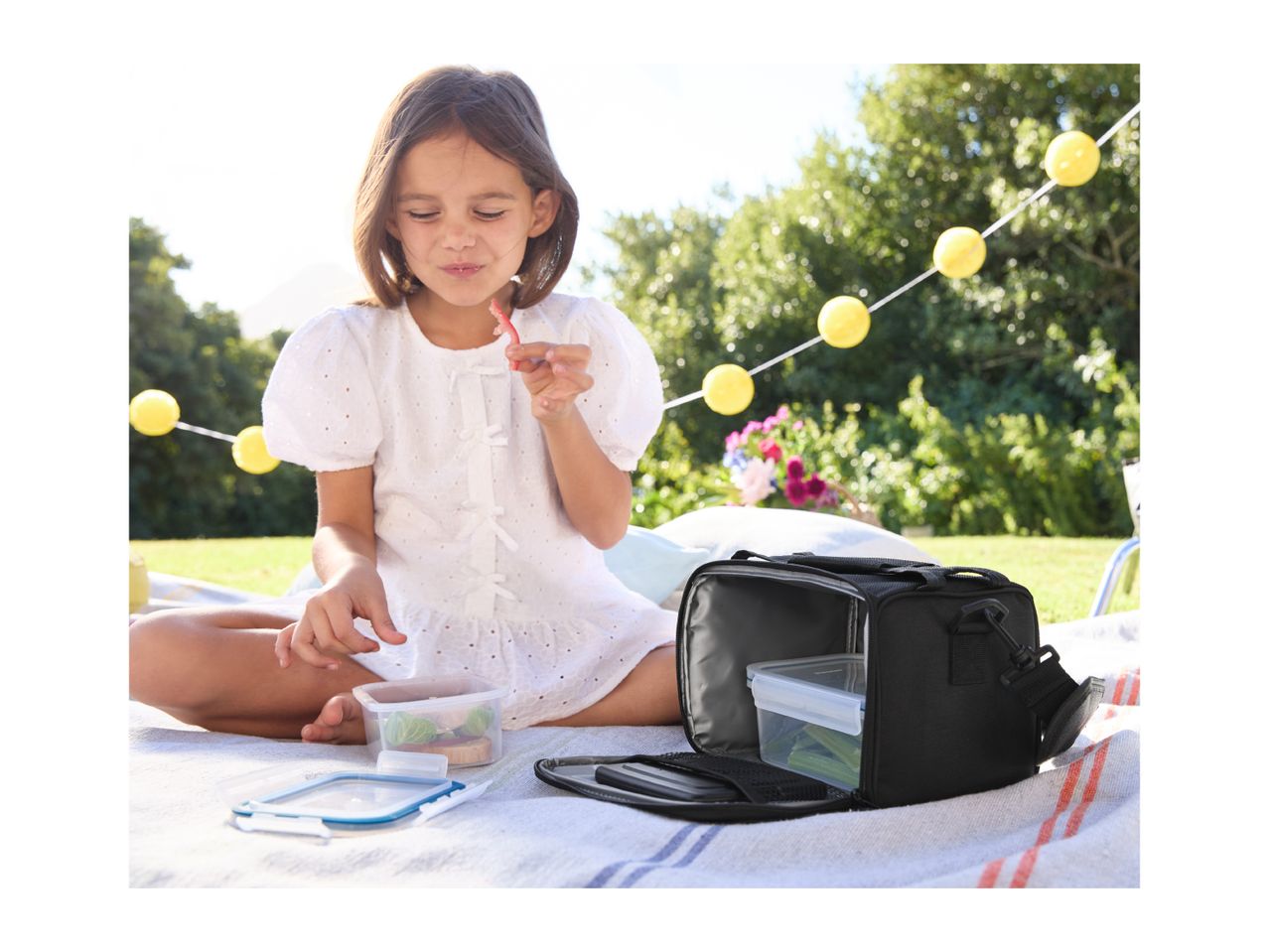 A girl enjoying a picnic with a lunch bag and food containers on a blanket.