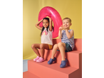 Two happy children in summer clothes and clogs, with a pink inflatable ring, sitting on coral steps.