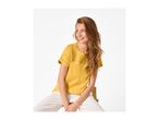 Woman in a yellow eyelet t-shirt and white trousers, sitting on a wooden stool.