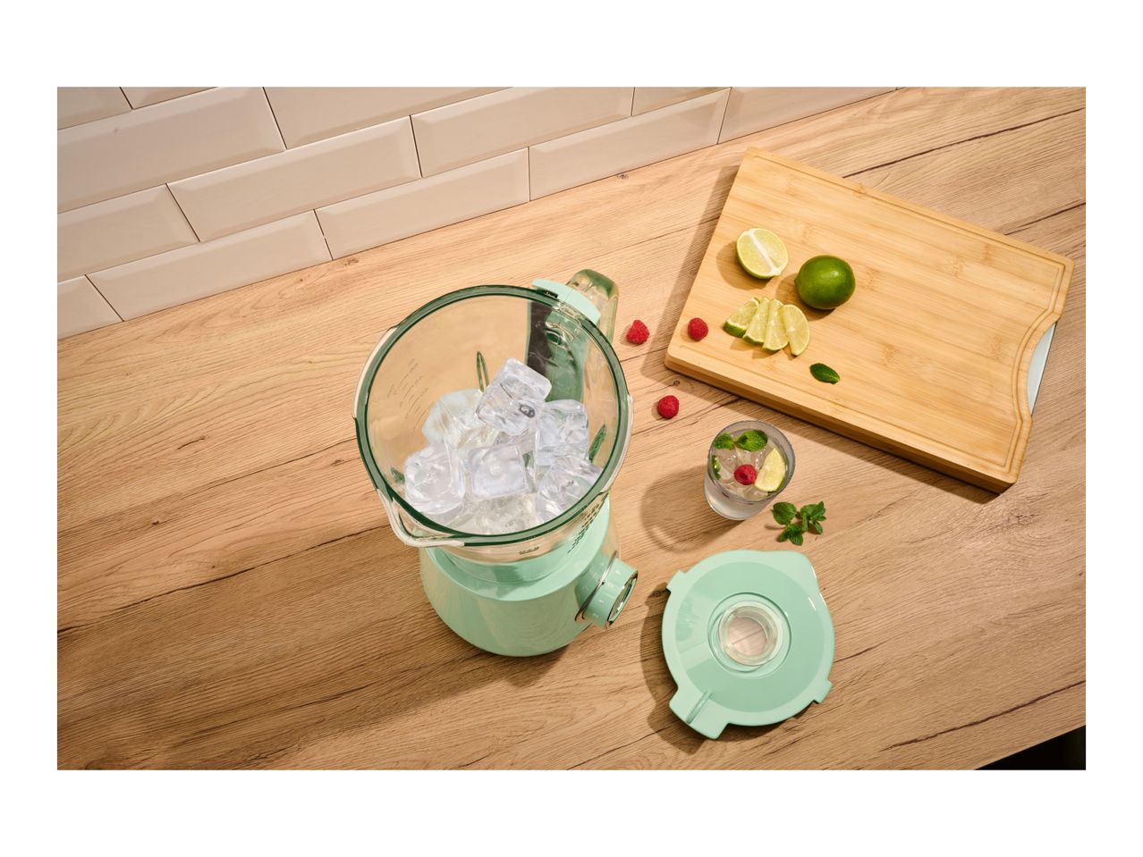 Blender with ice, limes, raspberries, and a drink on a wooden counter.