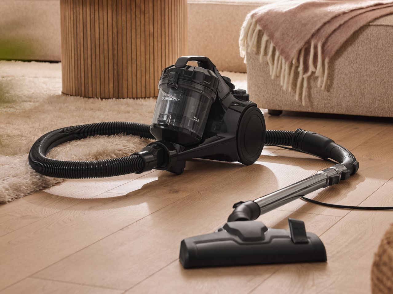 Black vacuum cleaner on a wooden floor next to a fluffy rug and sofa.