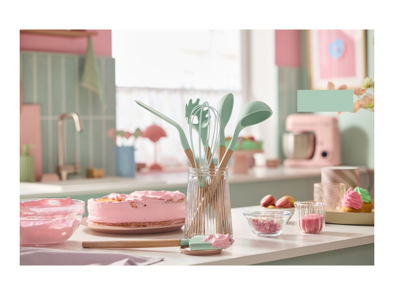 A kitchen scene with baking tools, a pink cake, and macarons on a white counter.