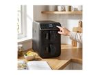 A person's hand operates a black dual-basket air fryer on a wooden kitchen counter with a cookbook and ingredients.