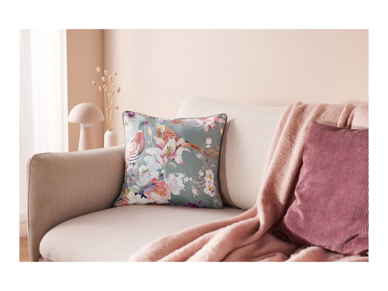 A light-colored sofa with a floral pillow, a pink throw blanket, and a ribbed pink pillow.