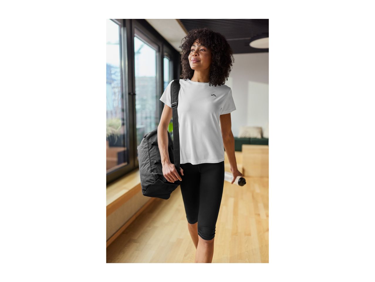 Woman in white sports t-shirt and black capri leggings, carrying a gym bag and water bottle.