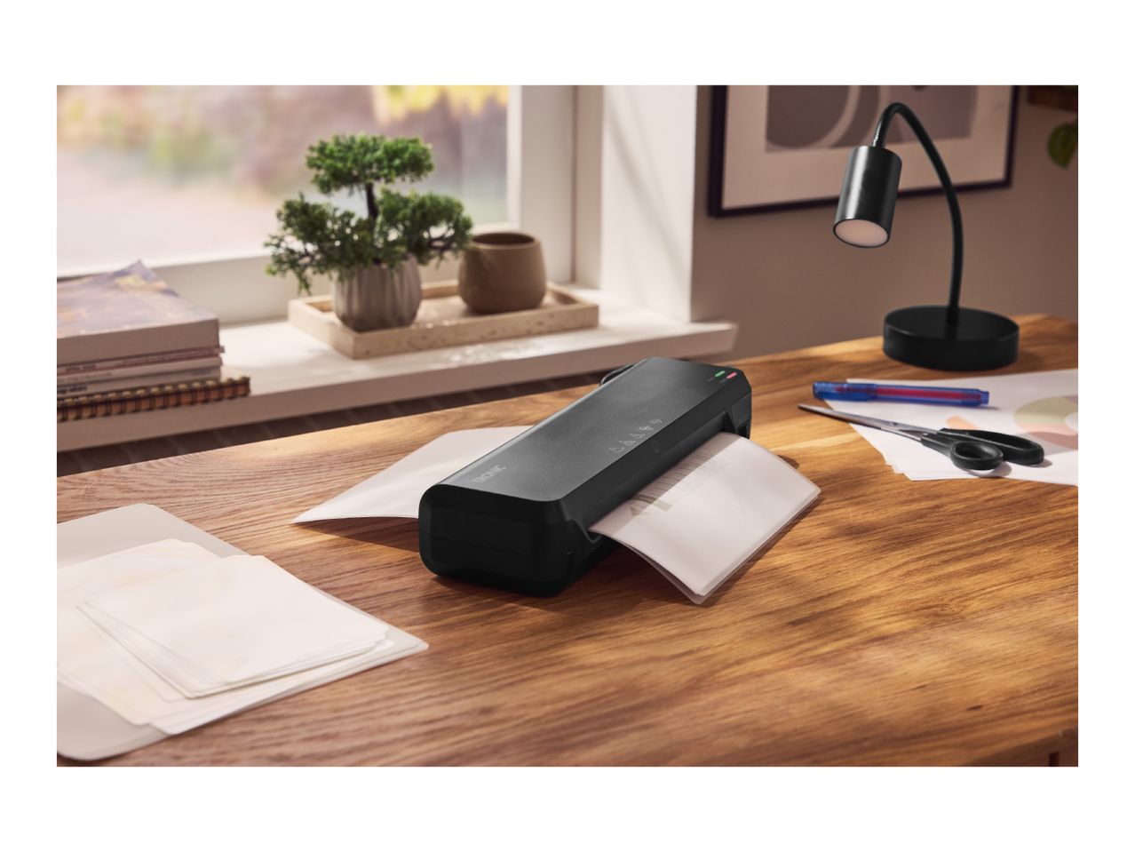 Black laminator on a wooden desk, laminating a document, with office supplies nearby.