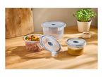 Three round food storage containers, one with pasta salad, one with soup, and one empty, on a wooden table.