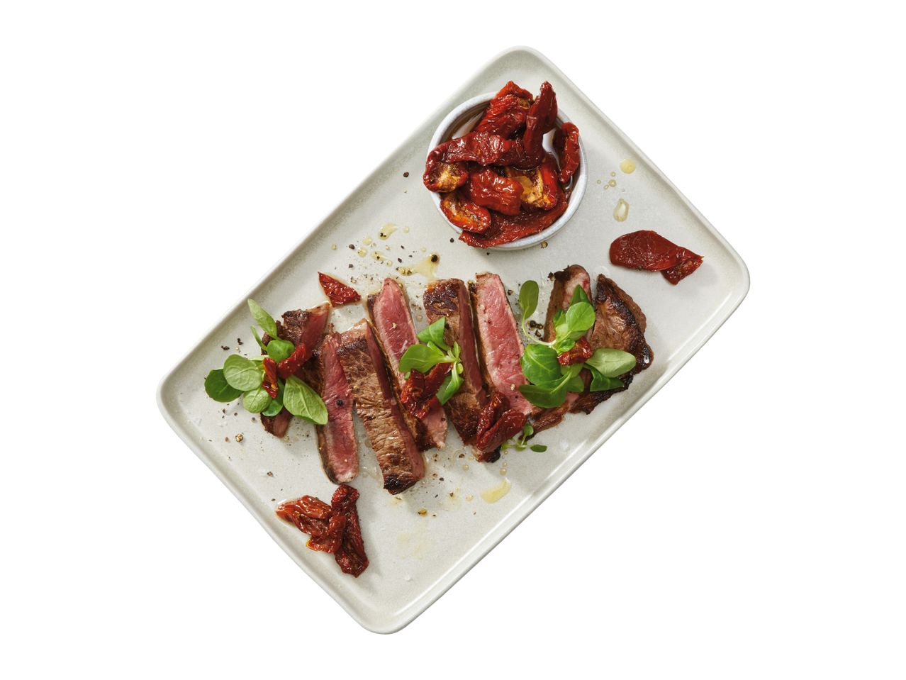 Sliced steak with sun-dried tomatoes and lamb's lettuce on a rectangular plate.