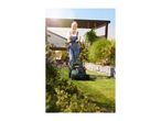 Smiling woman in overalls mowing a sunny lawn with a green lawnmower.