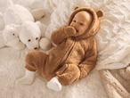 Baby in braunem Teddy-Overall auf flauschiger Decke mit weißen Plüschtieren.