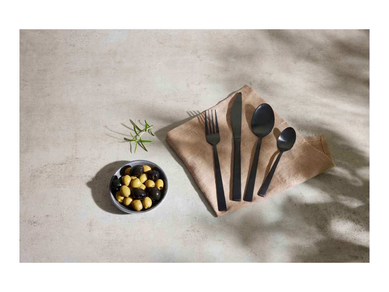 Black cutlery set on a beige napkin with a bowl of olives and rosemary sprig.
