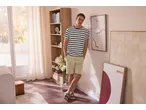 Bărbat în tricou în dungi și pantaloni scurți kaki, sprijinit de un perete cu artă abstractă.