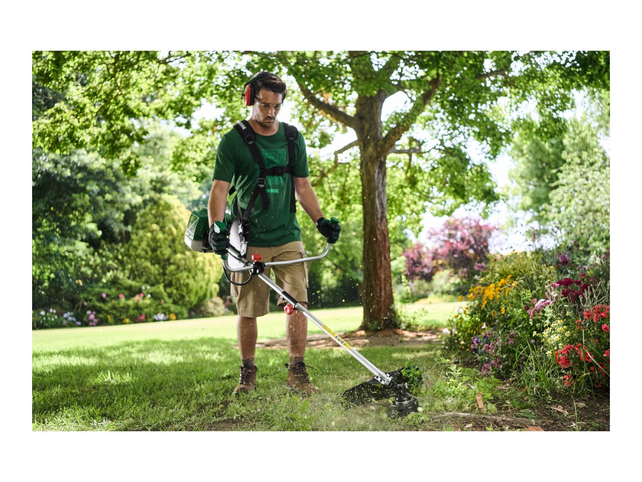 Man using a brush cutter in a garden, wearing safety glasses and ear protection.