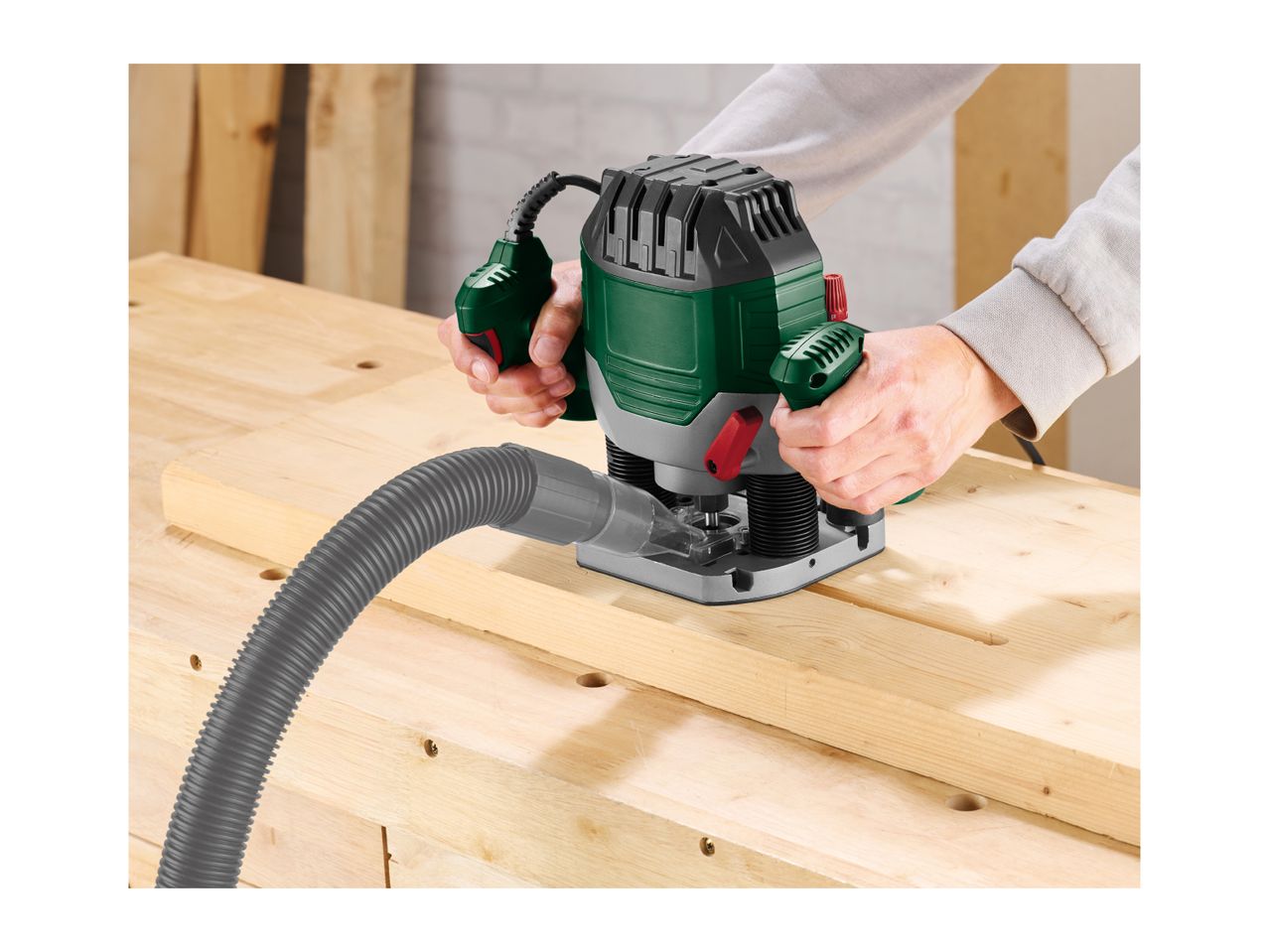 Person using a plunge router with a dust extraction hose on a wooden workbench.