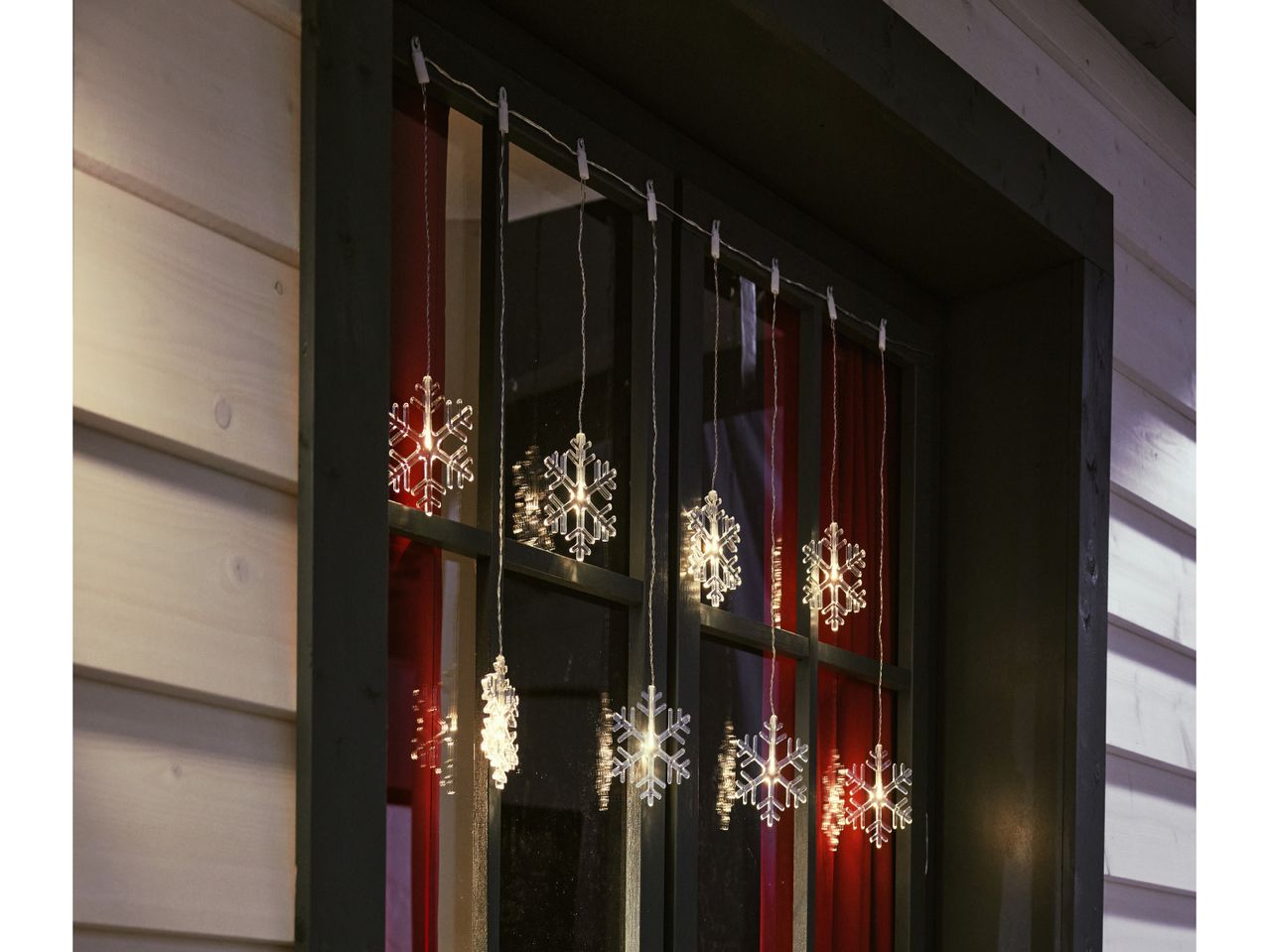 Snowflake string lights hanging in a window with red curtains, illuminating a wooden house.