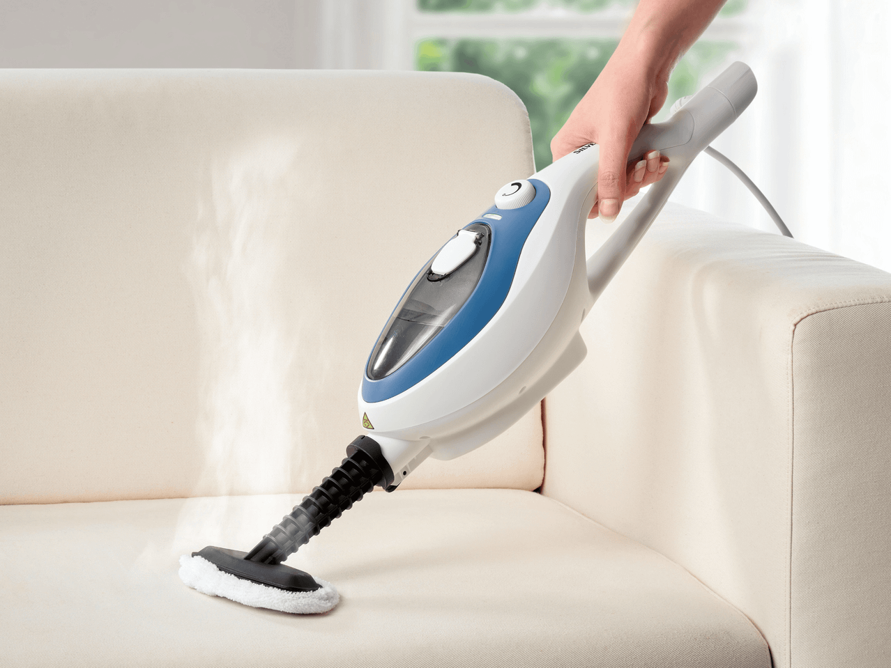 Eine Hand hält einen Polsterdampfreiniger und reinigt damit ein weißes Sofa.
