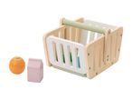 A wooden toy shopping basket with a green handle, next to a toy orange and a pink milk carton.