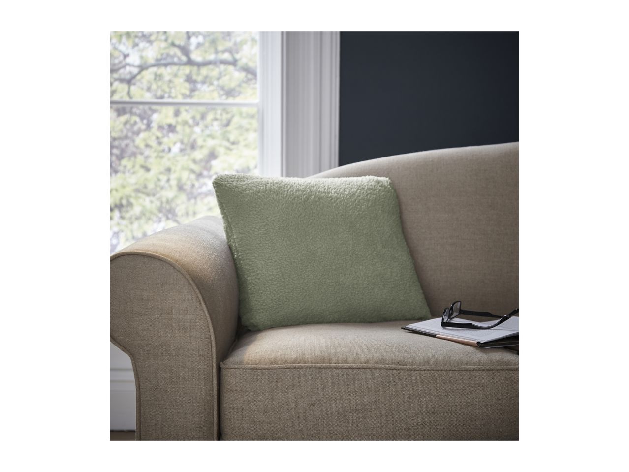 A cozy green cushion on a beige sofa with a notebook and glasses, next to a window.