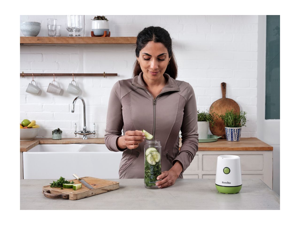 Woman adding cucumber to a blender bottle with greens, next to a personal blender.