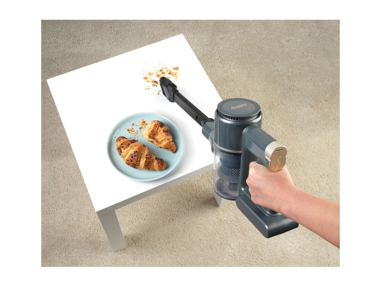 Handheld vacuum cleaning croissant crumbs off a white table next to a plate of croissants.
