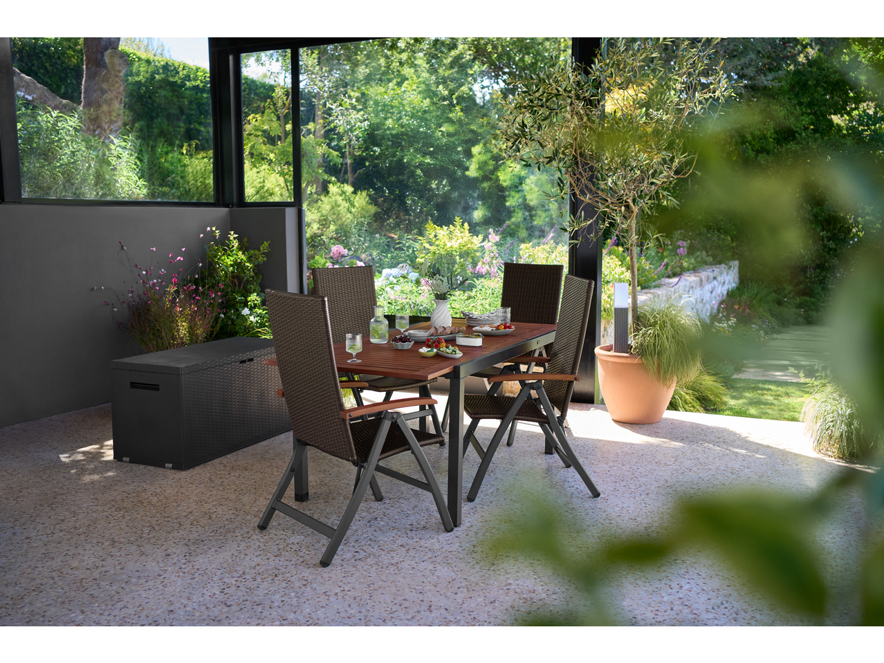 Outdoor dining set with a brown table and chairs, next to a black storage box, in a sunroom.
