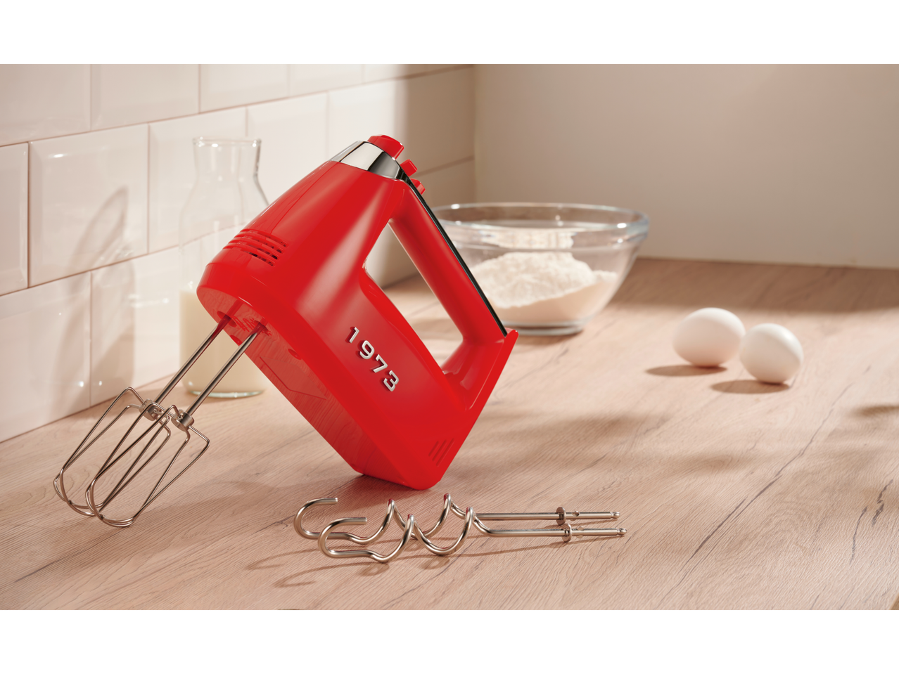 A red hand mixer with beaters and dough hooks on a wooden counter with baking ingredients.
