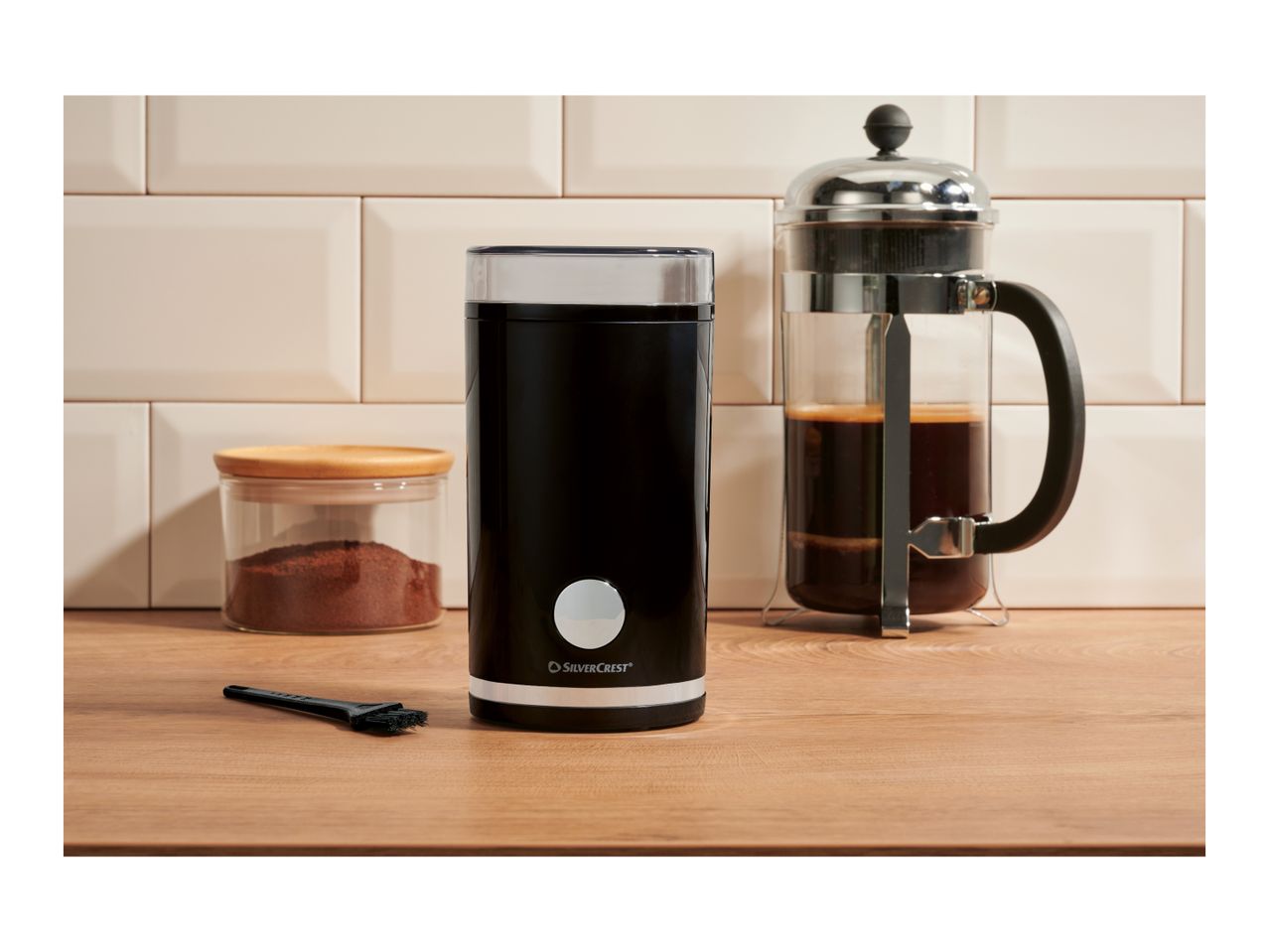Black coffee grinder, French press, and ground coffee jar on a wooden counter.