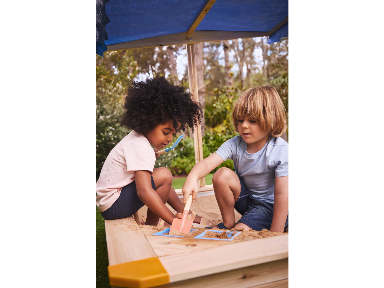Two children playing in a wooden sandpit with sand toys under a blue canopy.