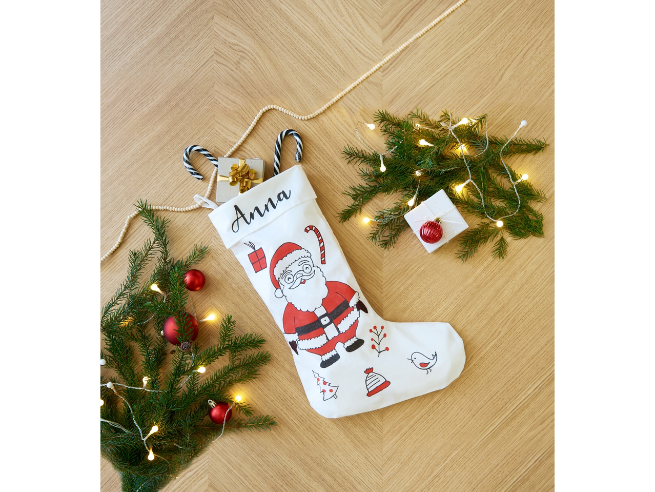 Personalizēta Ziemassvētku zeķe ar Ziemassvētku vecīti, dekorācijām un gaismiņām uz koka galda.
