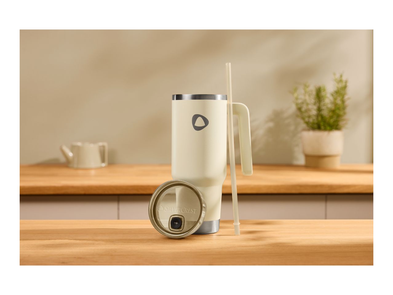 Cream-colored insulated tumbler with a handle and straw, next to its lid, on a wooden counter.