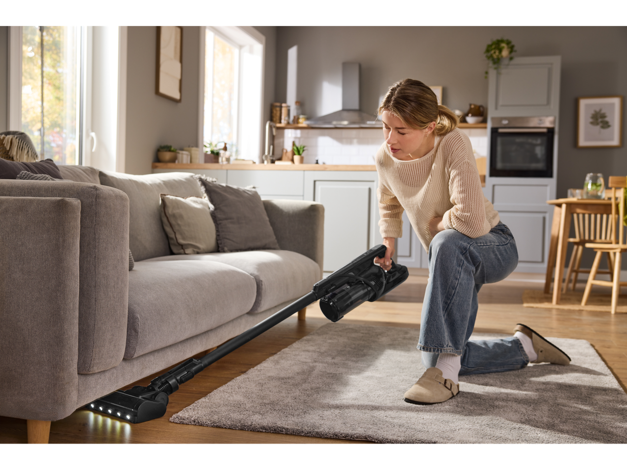 Mulher a aspirar debaixo do sofá com o aspirador vertical sem fios 2 em 1 Silvercrest®.