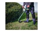 A person using a cordless grass trimmer to cut grass next to a hedge.