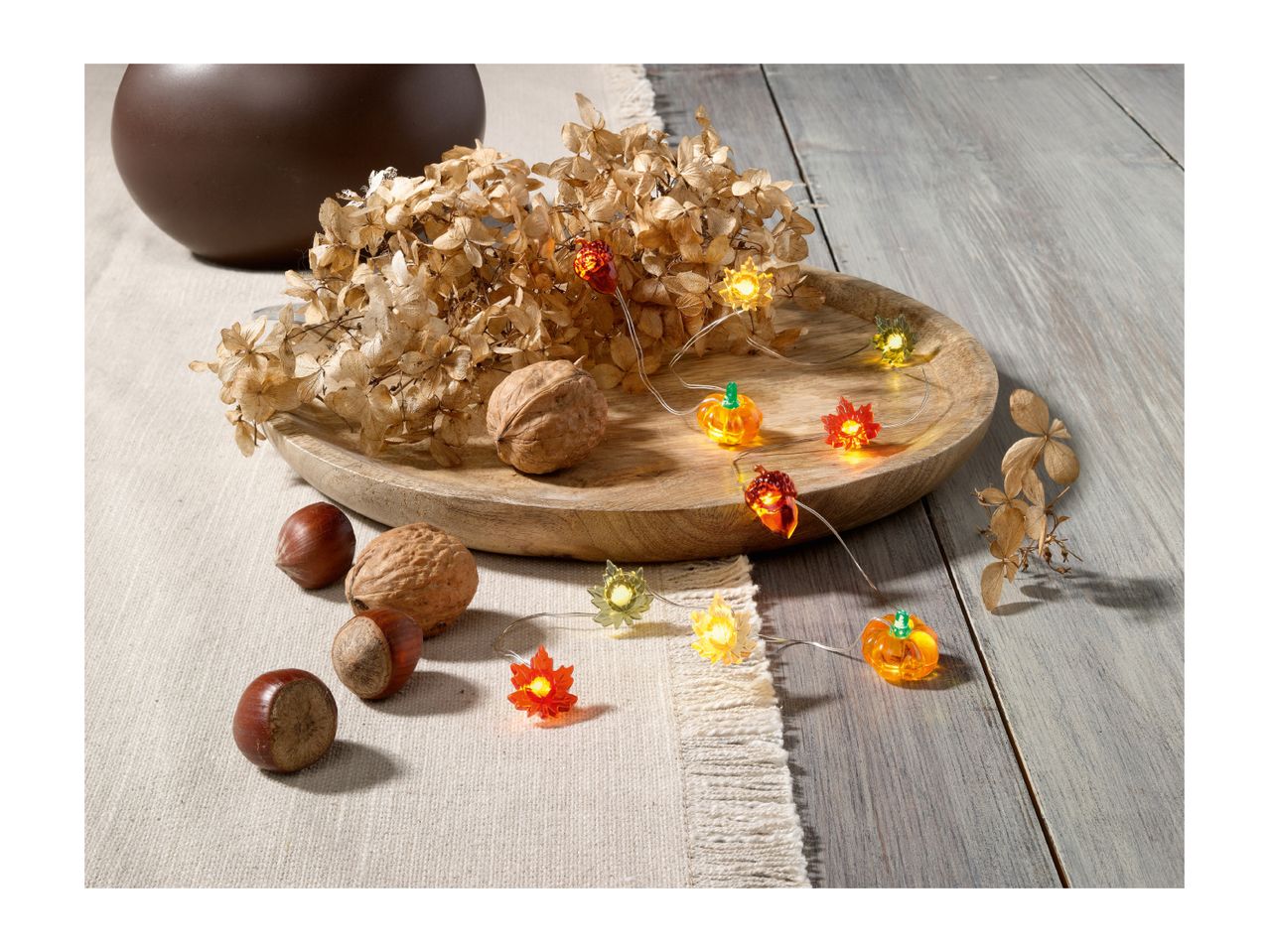 Autumnal string lights with pumpkins and leaves, dried flowers, and nuts on a wooden tray.