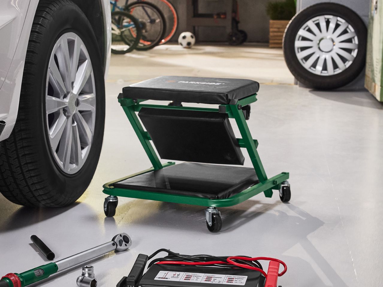 Green workshop creeper with tools and car battery charger next to a car wheel.