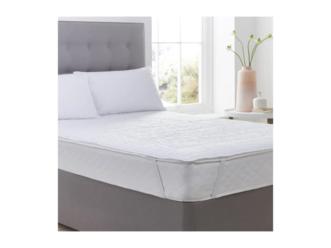 A bed with a grey upholstered headboard and base, featuring a white quilted mattress protector.