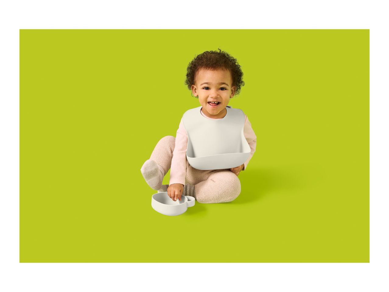 Happy baby in a bib, holding a bowl and spoon, ready to eat.
