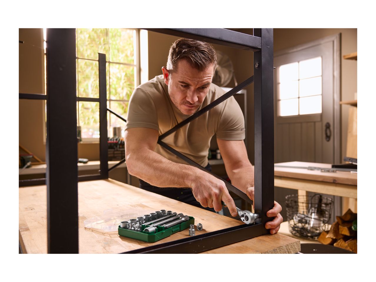Man assembling a metal frame with a ratchet wrench and socket set on a wooden workbench.