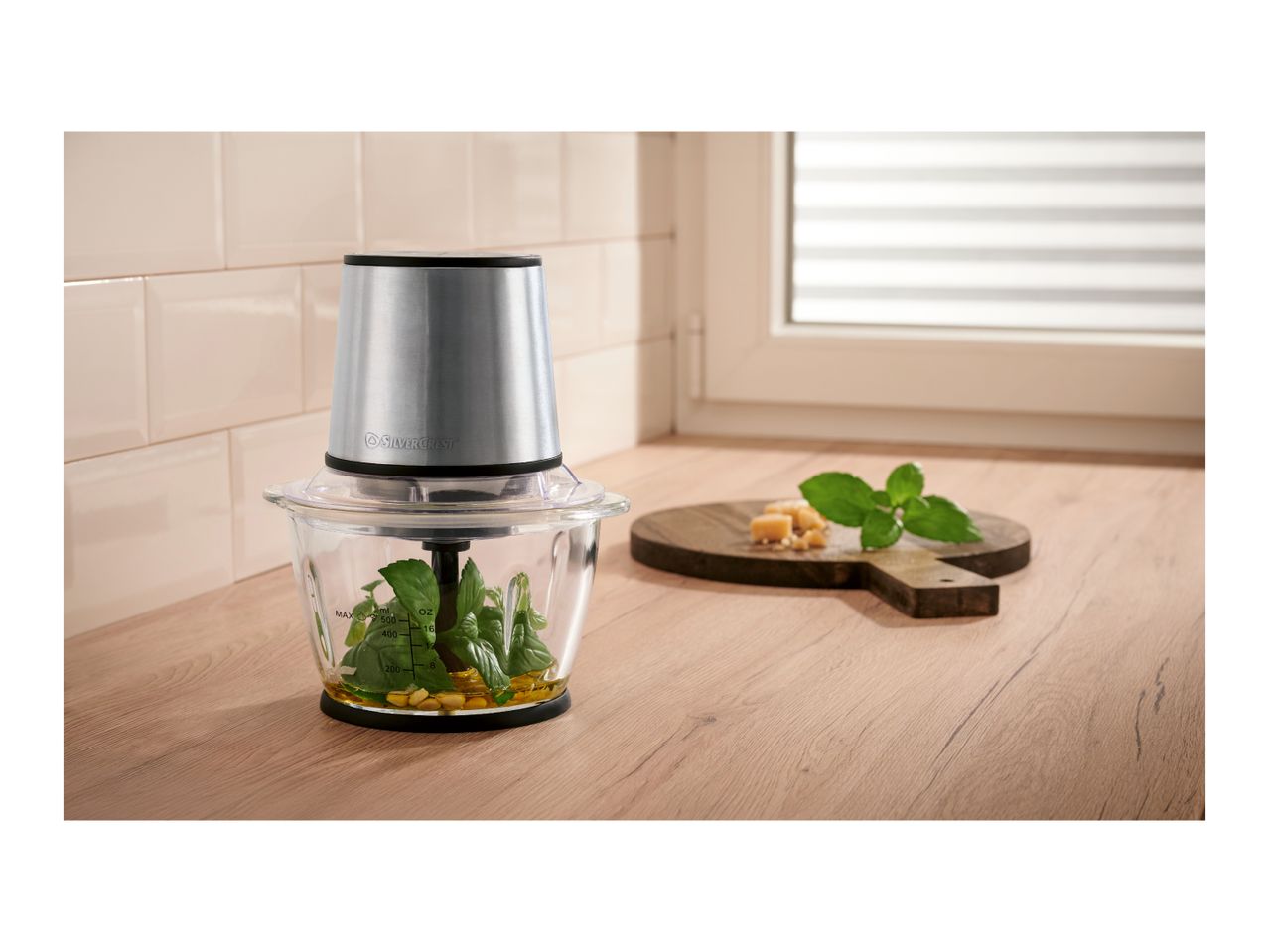 Food chopper with basil, pine nuts, and oil, next to a wooden board with cheese and mint.