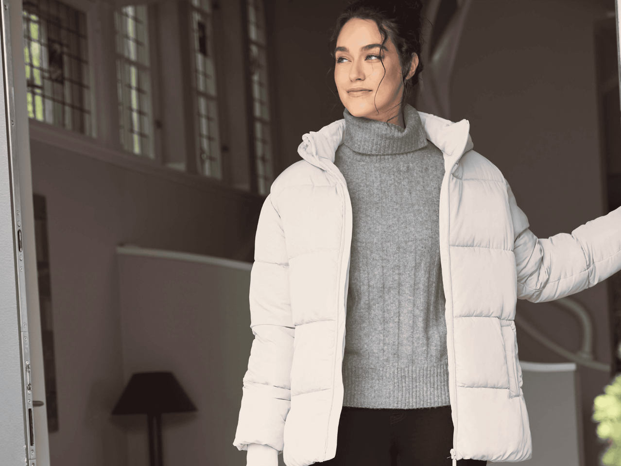 Frau in beiger Pufferjacke und grauem Rollkragenpullover, die aus einer Tür schaut.
