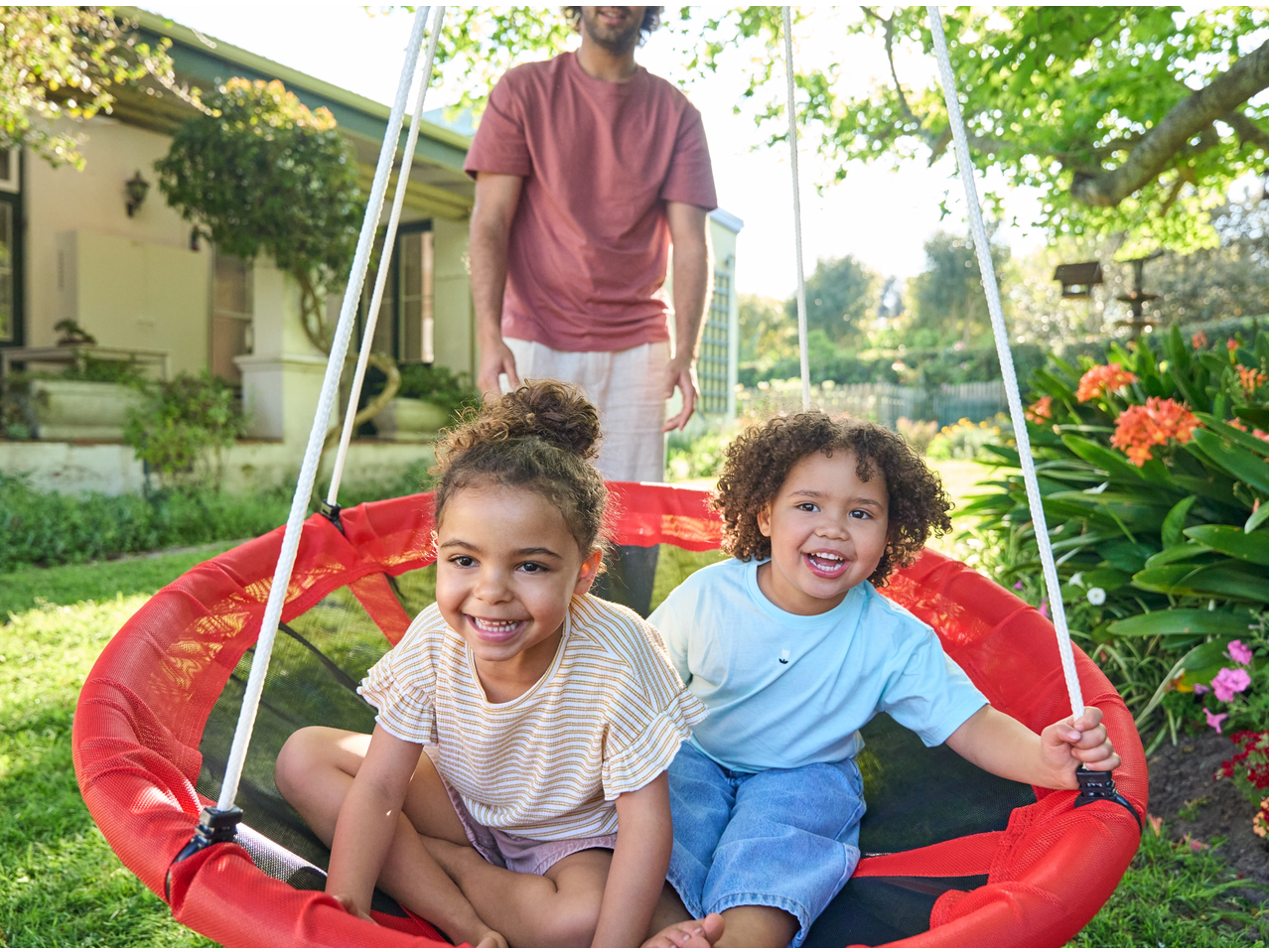 Two happy children in a red swing, with an adult standing behind them in a garden.