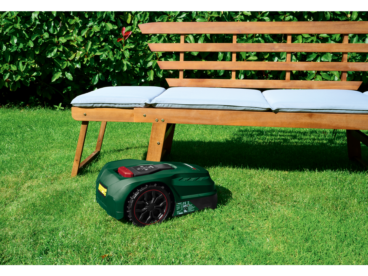 Robotic lawnmower on green grass next to a wooden garden bench with light blue cushions.