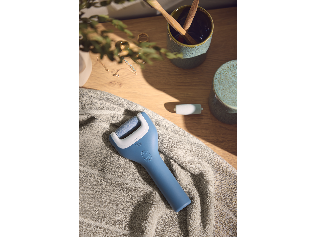 Lixa elétrica para pés CIEN BEAUTY sobre uma toalha, com escovas de dentes de bambu e joias ao fundo.