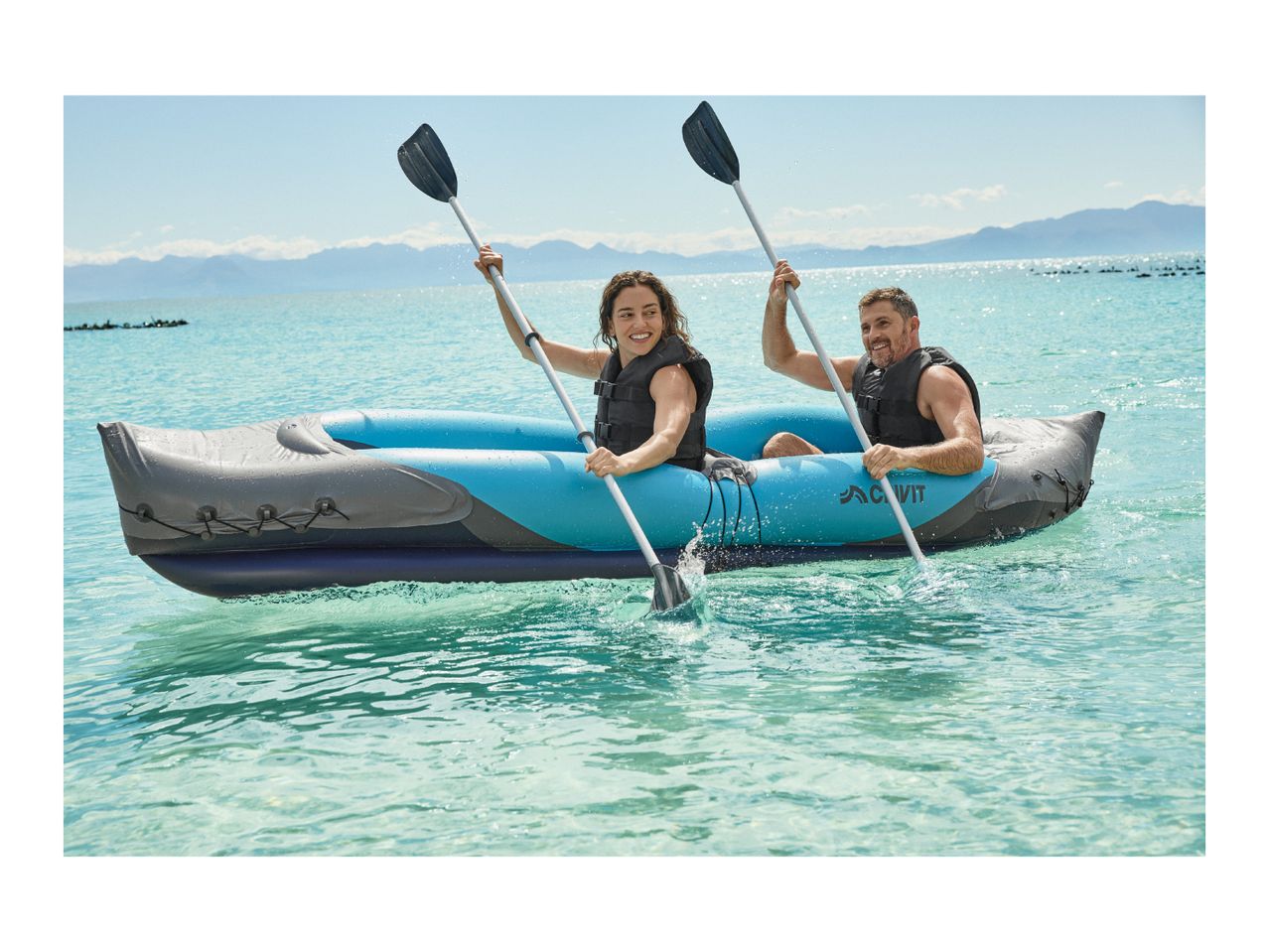 Two people in an inflatable kayak on clear blue water, paddling and smiling.