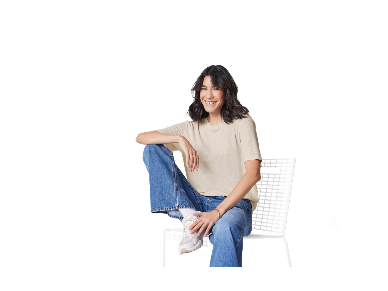 Mulher sorridente sentada em uma cadeira branca usando jeans e tênis.