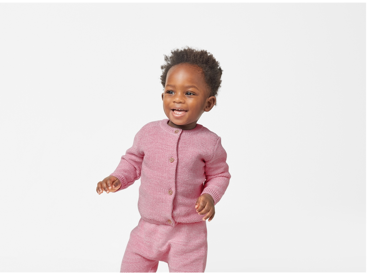 A smiling baby in a pink knitted cardigan and matching pants.