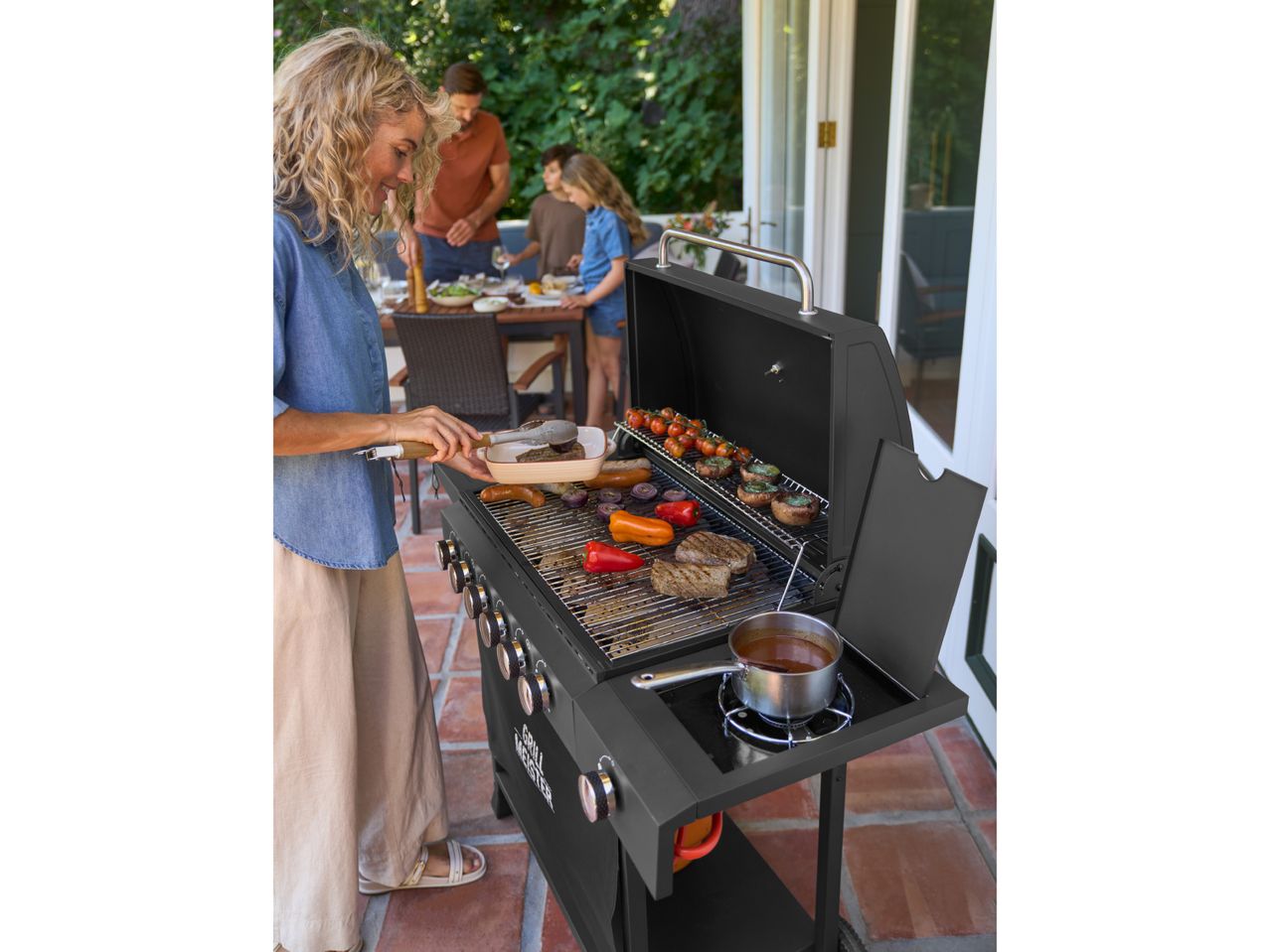 Woman grilling meat and vegetables on a barbecue, with family dining in the background.