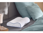 Ergonomic pillow on a bed with blue bedding and a lamp on a nightstand.