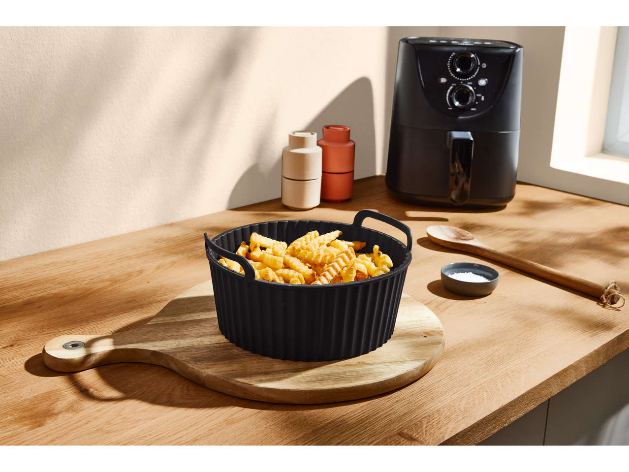 Cartofi prăjiți crocanți într-un vas negru de silicon, cu un air fryer în fundal.
