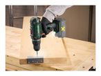 A person using a cordless drill to attach a metal hinge to a wooden board on a workbench.