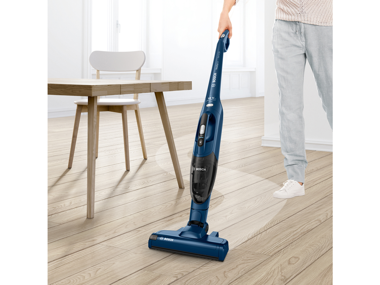 Person vacuuming light wooden floor with a blue cordless stick vacuum cleaner in a bright room.